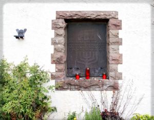Die dunkle Metallplatte ist in eine weiße Mauer eingelassen und wird von Bruchsteinen umrahmt. Die Inschrift der Gedenktafel lautet: Hier stand bis zur Zerstörung durch die Nationalsozialisten am 9.11.1938 die Synagoge der Jüdischen Gemeinde Honnef“. Auf dem Mauervorsprung vor der Tafel stehen drei rote Friedhofskerzen.