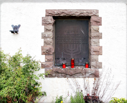 Die dunkle Metallplatte ist in eine weiße Mauer eingelassen und wird von Bruchsteinen umrahmt. Die Inschrift der Gedenktafel lautet: Hier stand bis zur Zerstörung durch die Nationalsozialisten am 9.11.1938 die Synagoge der Jüdischen Gemeinde Honnef“. Auf dem Mauervorsprung vor der Tafel stehen drei rote Friedhofskerzen.