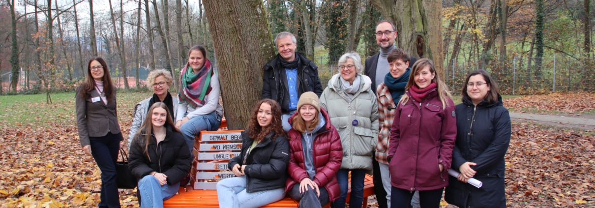 Park mit Baum und orange Bank: Schülerinnen, sitzend und Personen stehend