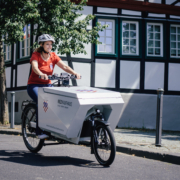 Eine Frau fährt auf einem Lastenrad über die Straße. Sie trägt eine blaue Jeans, ein rotes T-Shirt und einen weißen Fahrradhelm auf dem Kopf. Die Ladebox des Lastenrads ist mit der Bad Honnefer Stadtmarke sowie dem Kiezkaufhaus-Logo gekennzeichnet.