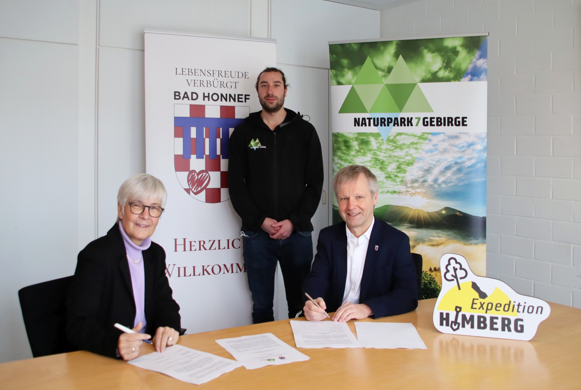 Zwei Personen - Brigitte Kohlhaas und Bürgermeister Otto Neuhoff, sitzend und eine Person - Linus Steinbach - stehend mit Werbetafel für naturpark Siebengebirge