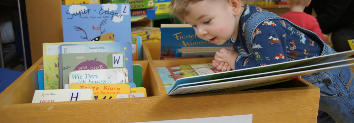 Ein Kleinkind beugt sich über eine Kiste mit Bilderbüchern und schaut vertieft auf die Seiten eines großen Buches.