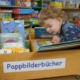 Ein Kleinkind beugt sich über eine Kiste mit Bilderbüchern und schaut vertieft auf die Seiten eines großen Buches.