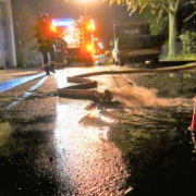 Es ist dunkle Nacht. Auf einer beleuchteten Straße liegen Feuerwehrschläuche.Aus überfluteten Kellnern abgepumptes Regenwasser sprudelt über den Asphalt.