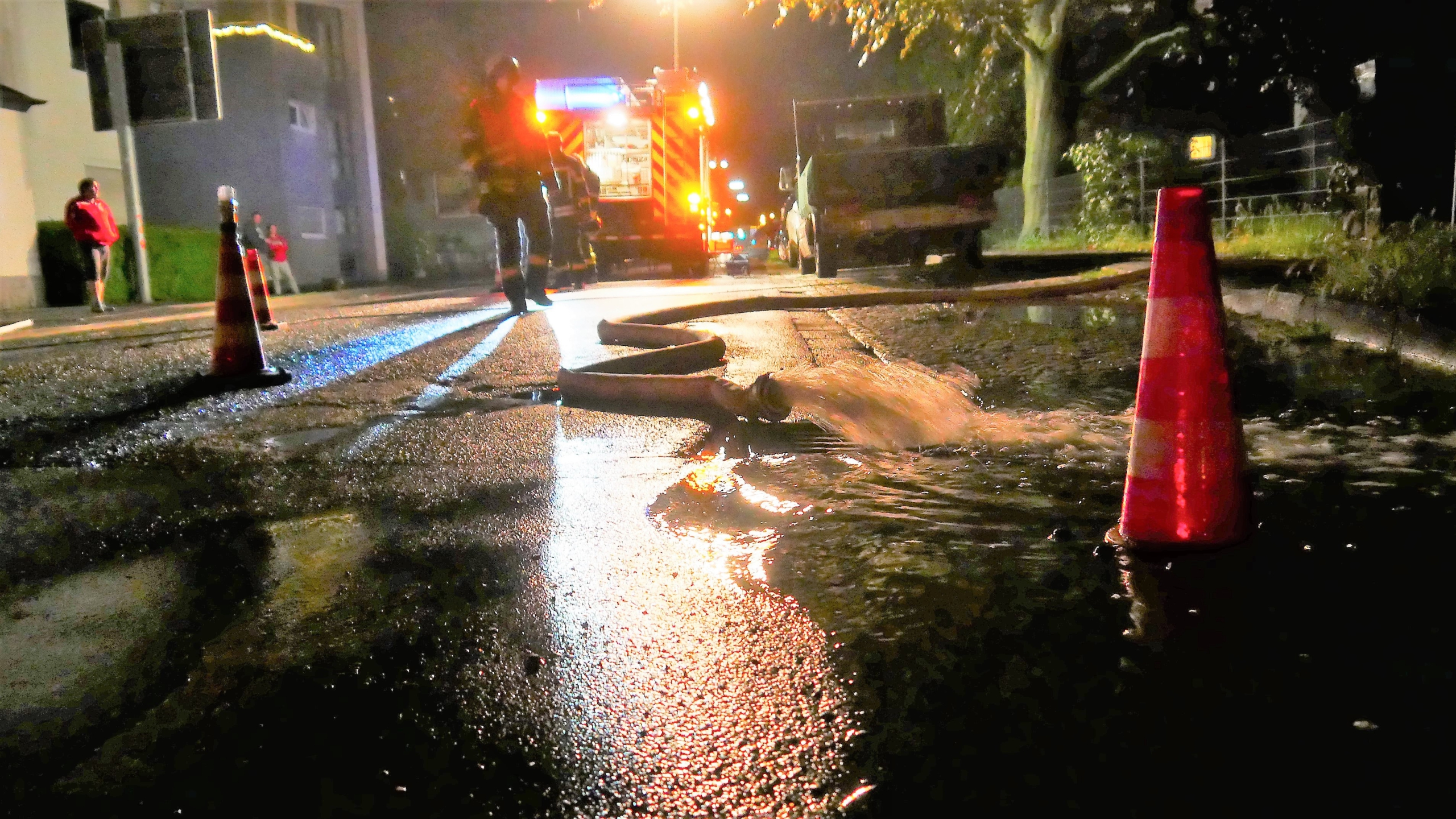 Es ist dunkle Nacht. Auf einer beleuchteten Straße liegen Feuerwehrschläuche.Aus überfluteten Kellnern abgepumptes Regenwasser sprudelt über den Asphalt.