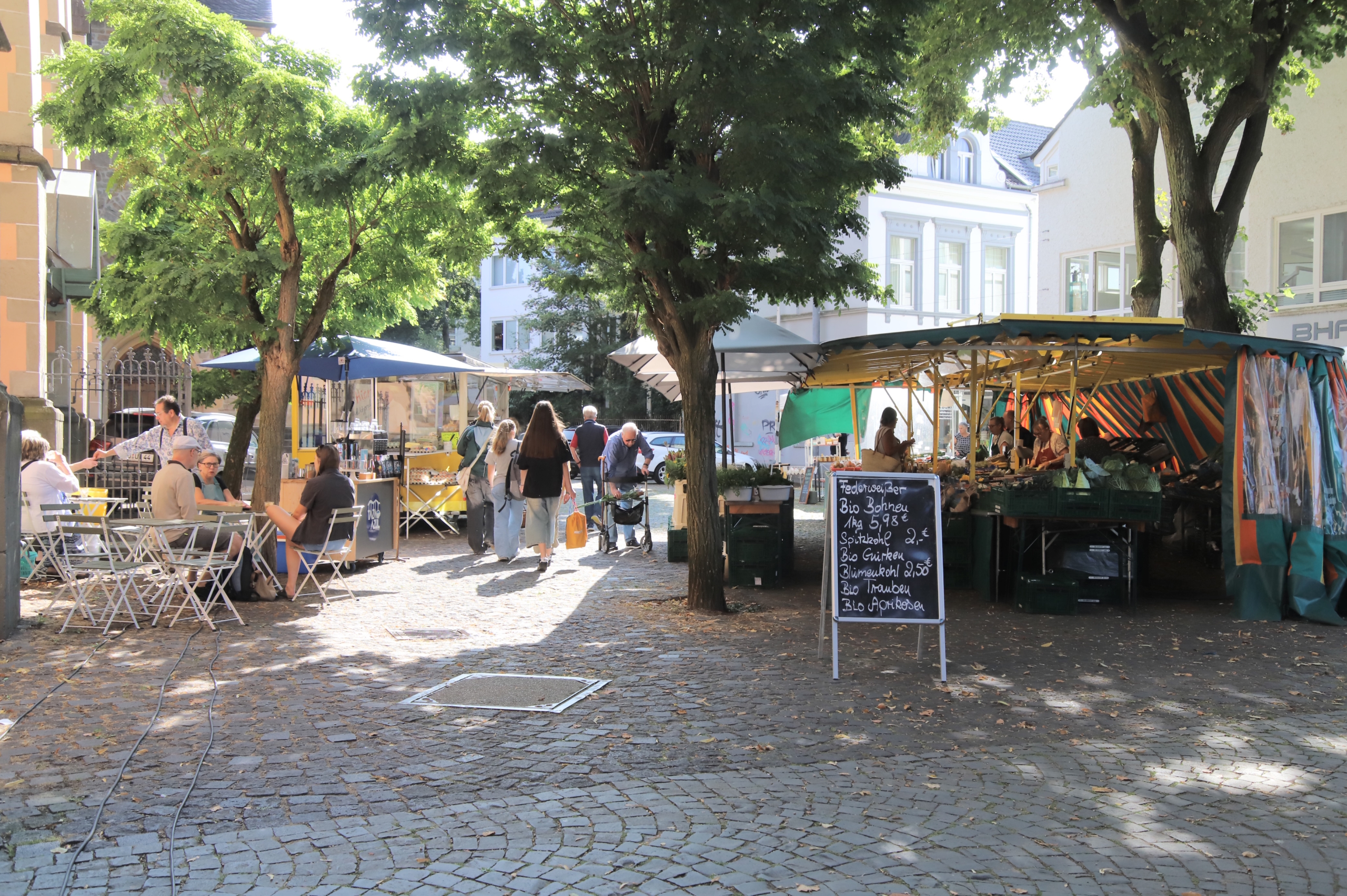Besucher schlendern über den Wochenmarkt auf dem Kirchplatz in Bad Honnef. Links sitzen Menschen an einem Café unter Bäumen, rechts bieten Händler regionale Bio-Produkte unter einem Zelt an.